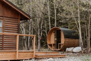 Metsän keskellä sijaitseva hirsihökkelö, jossa on puinen terassi ja suuri sylinterinmuotoinen rakennelma, todennäköisesti sauna.
