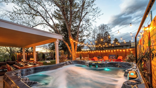 Un jacuzzi con terraza de madera y luces colgantes, rodeado de mobiliario exterior y una zona cubierta.