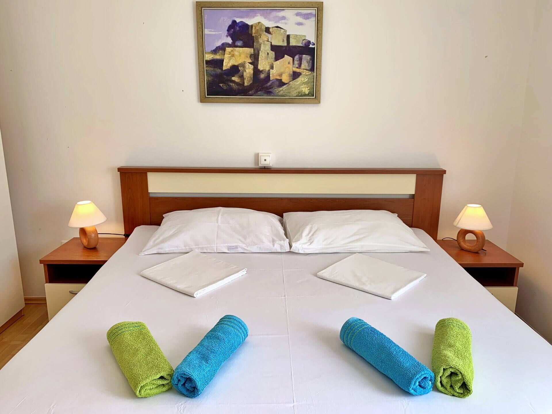 A neatly made bed with white linens, two pillows, and two rolled towels in green and blue. There are two bedside lamps on wooden nightstands, and a framed painting hangs on the wall above the bed.