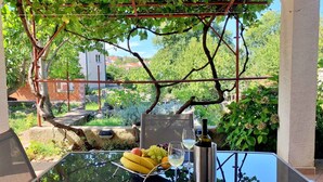 Une table d’extérieur avec un plateau en verre, entourée d’une pergola couverte de plantes grimpantes, d’une bouteille et d’un panier de fruits.
