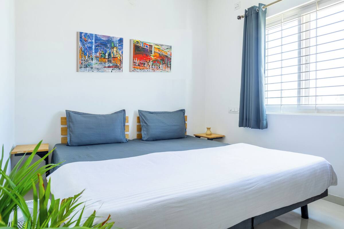 Un dormitorio con una cama, dos almohadas, una mesita de noche, un jarrón y dos cuadros en la pared.