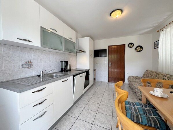 Una cocina compacta con armarios blancos, una estufa integrada y una cafetera. Hay una zona de comedor con una mesa de madera y un sofá.