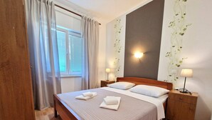 A neatly made bed with pillows and folded towels, a bedside table with a lamp, a wooden nightstand, and a window with curtains.