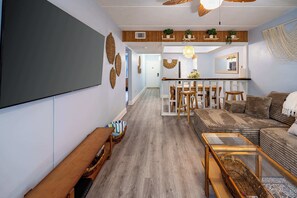 A modern living room with a sofa, coffee table, and TV. There is a dining area with a table and chairs. The room has wooden flooring and a ceiling fan.