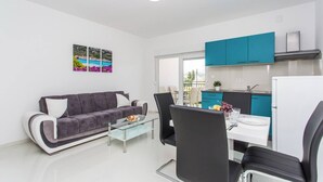 A modern living room with a grey sofa, a glass coffee table, and a dining area with a white table and black chairs. There is a kitchen with teal cabinets and a white refrigerator.