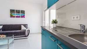 A modern kitchen with teal cabinets, a stainless steel sink, and a glass coffee table with pastries. There is a sofa with a cushion and a plant on the counter.