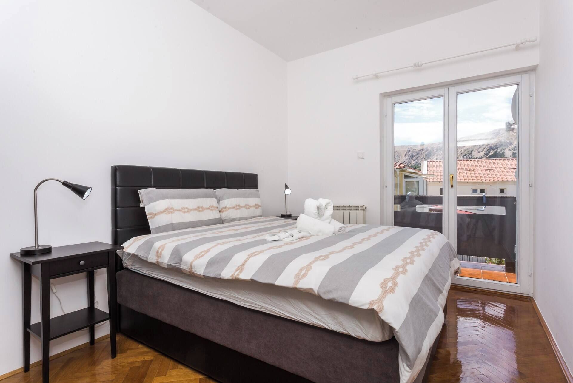 A bedroom with a bed, a nightstand, a lamp, and a view of a balcony.