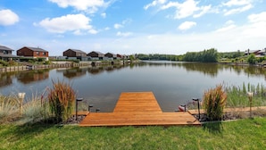 Una vista serena de un lago con un muelle de madera que se adentra en aguas tranquilas, rodeado de casas modernas y exuberante vegetación.