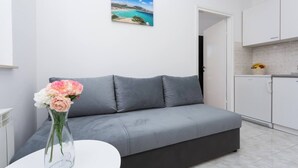 Un salon moderne avec un canapé gris, une petite table blanche et un vase rempli de fleurs. Un tableau représentant un paysage côtier est accroché au mur.