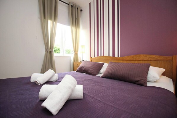 A neatly made bed with purple bedding and white towels, a wooden headboard, and a striped wall in the background.