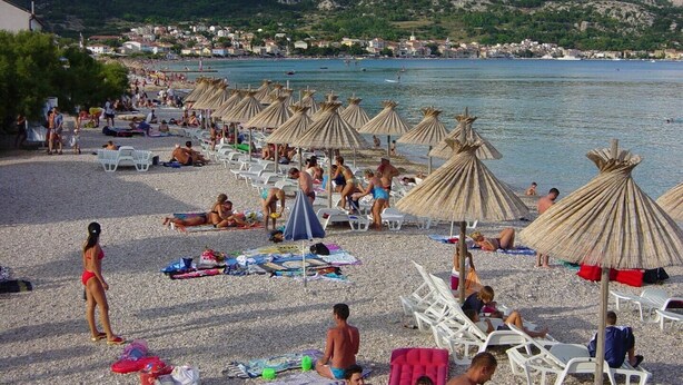 Une plage bordée de rangées de chaises longues et d’ombrelles en chaume, une plage de galets, et une ville en arrière-plan.