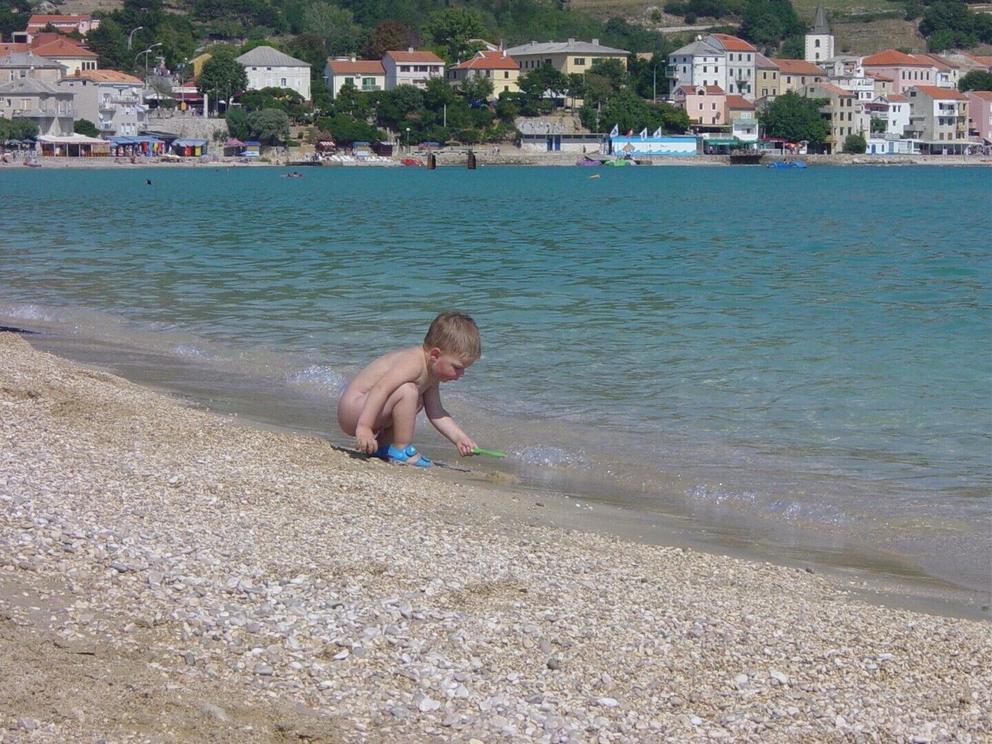 A child playing on a pebble beach with a body of water and buildings in the background.