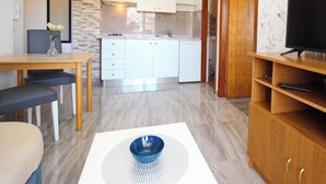 A compact kitchen with white cabinets, a dining area with a table and chairs, and a TV on a wooden stand. - Charming Apartment Brnic No. 4 - with spacious Balcony (Baska)