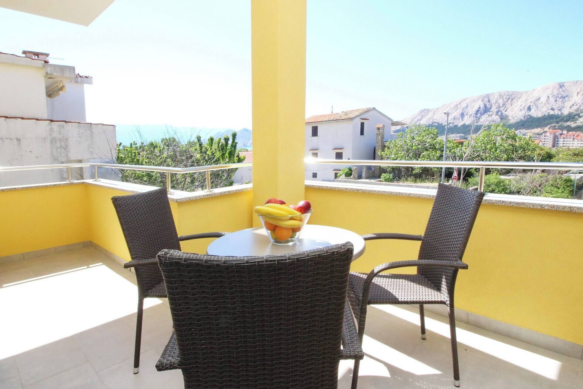 A balcony with two wicker chairs and a small round table, a bowl of fruit, and a view of buildings and mountains in the background.