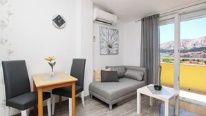 En moderne stue med en grå sofa, et lille bord og to stole. Der er et billede på væggen og en aircondition.