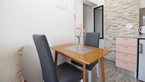 Un petit coin repas avec une table en bois, deux chaises, un vase avec une fleur, et un espace cuisine avec des placards roses et un réfrigérateur.