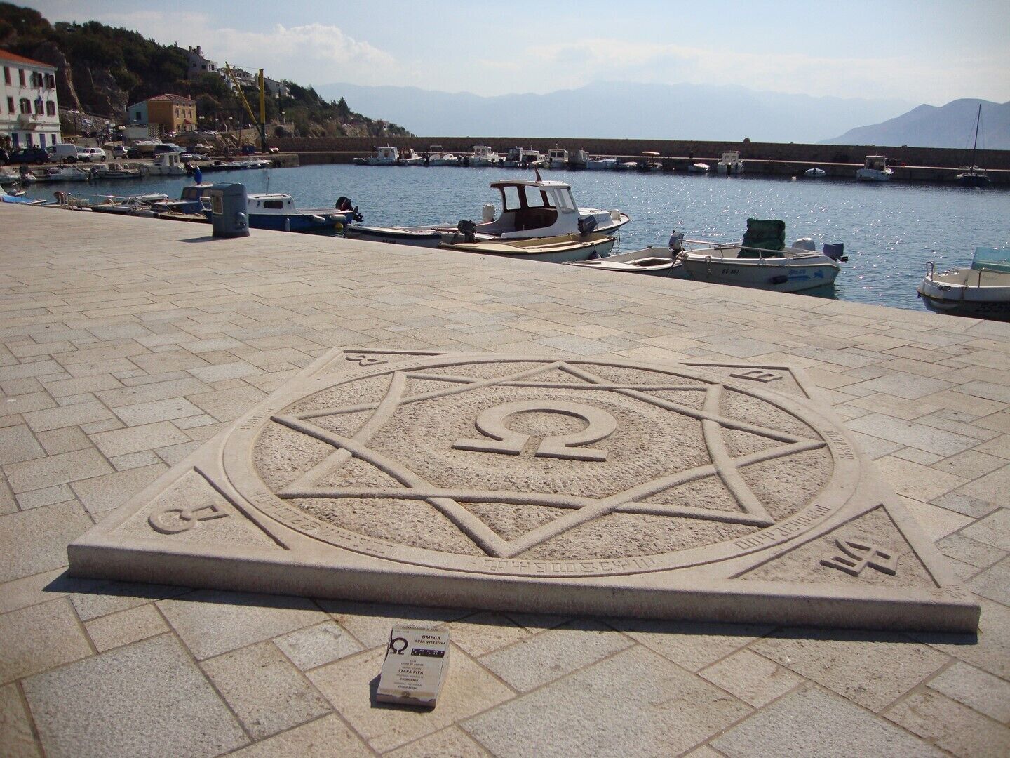 A paved area with a geometric pattern, boats docked in the background, and a clear sky.