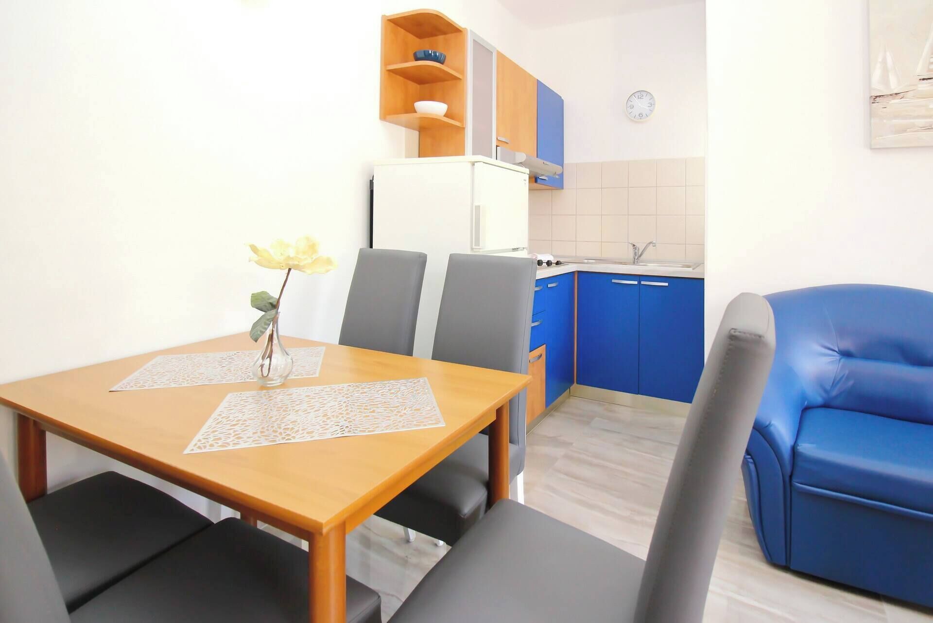 Een kleine keuken met blauwe kastjes, een witte koelkast en een eettafel met twee stoelen.