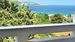 Un balcon avec un fauteuil en osier et une table, donnant sur un paysage côtier avec des arbres et une vue sur la mer.