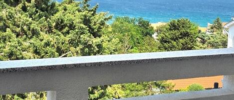 A balcony with a wicker chair and a table, overlooking a coastal landscape with trees and a view of the sea.