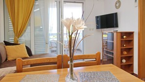 Un salon avec une table en bois, un vase rempli de fleurs blanches, un canapé avec des coussins jaunes, un téléviseur fixé au mur et une bibliothèque en bois.