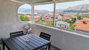 Uma varanda com mesa e cadeiras, dando vista para uma cidade costeira com edifícios e montanhas ao fundo.