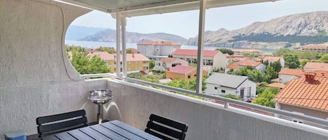 A balcony with a table and chairs, overlooking a coastal town with buildings and mountains in the background.