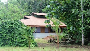 A house surrounded by dense greenery, with a sloped roof and a small garden area. - For 5 persons - 300 m from the beach in a tropical garden with biotope. (Cahuita)