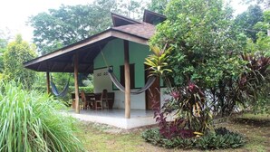 A small, single-story building with a covered patio area, surrounded by lush greenery and various plants. - For 5 persons - 300 m from the beach in a tropical garden with biotope. (Cahuita)