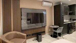A modern hotel room with a large bed, a TV mounted on the wall, a small dining table with a chair, a kitchenette, and a built-in air conditioning unit. - Meraki Homes at Loxwood House (Accra)
