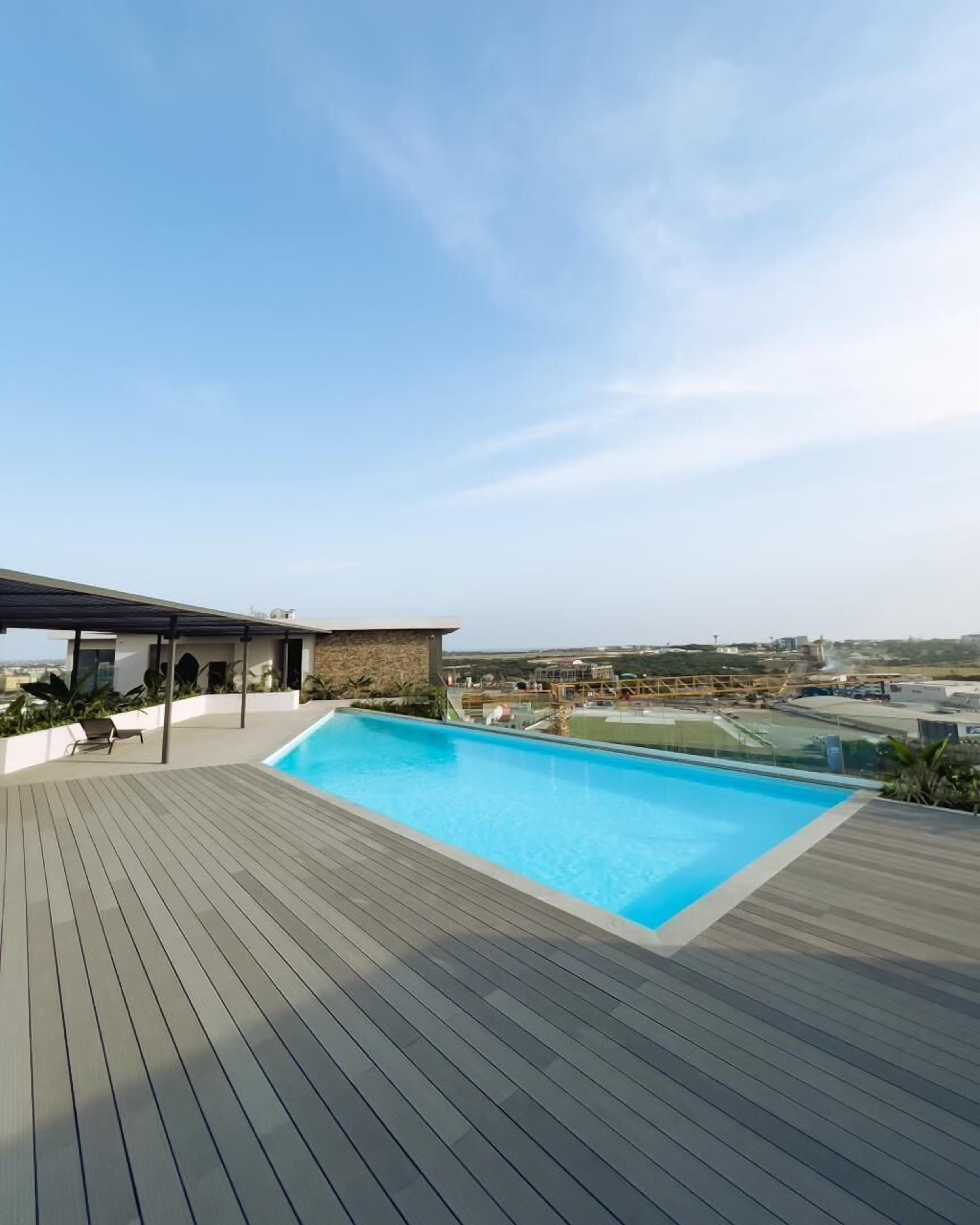 A rooftop swimming pool with a wooden deck, a covered seating area, and a view of the cityscape.