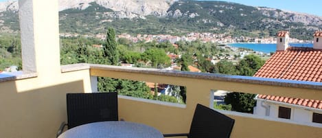 A balcony with a round table and two chairs, overlooking a coastal town with a mountainous backdrop.