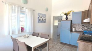 Eine kleine Küche mit blauen Schränken, einem weißen Esstisch und einem Fenster mit weißen Vorhängen.