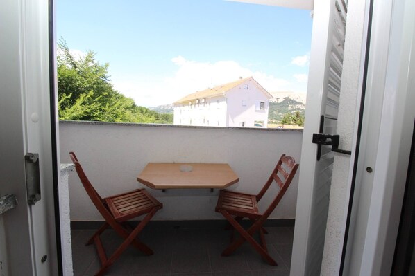 A balcony with a table and two wooden chairs, a white wall, and a view of buildings and trees.