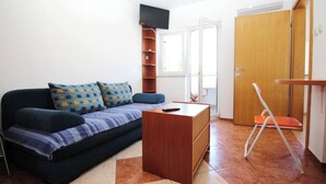 Een kleine, goed verlichte kamer met een bank, een houten bijzettafeltje, een stoel en een aan de muur bevestigde televisie.