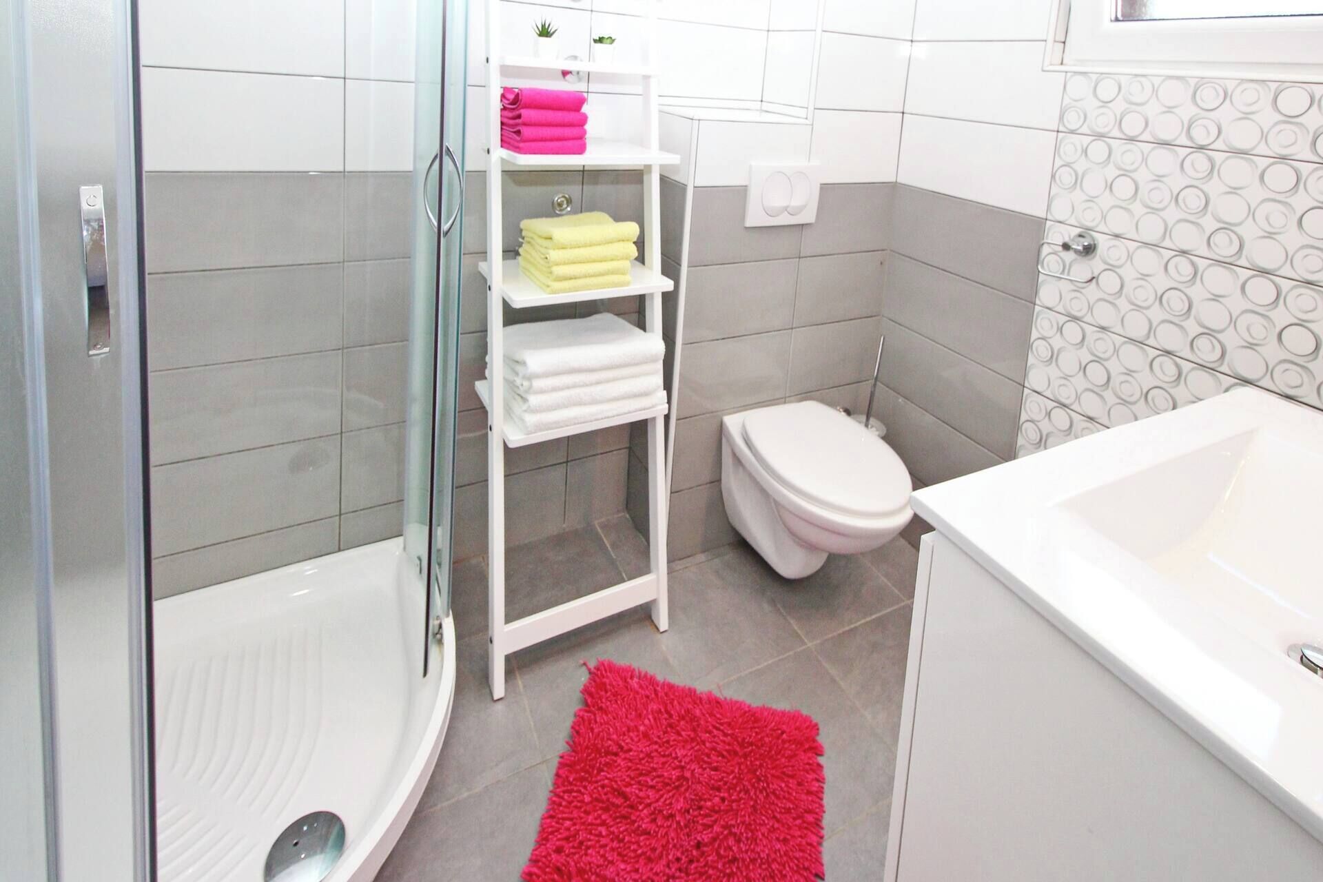 A modern bathroom with a glass shower enclosure, a white toilet, a white bathtub, and a shelf with folded towels.