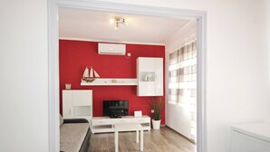 A compact living space with a red accent wall, white cabinetry, a flat-screen TV, a dining table, and a small plant. - Apartment Mihaela - 350m from the Beach (Baska)