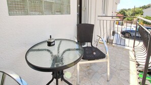 A small balcony with a glass table and a chair, a window with blinds, and a view of a parked car and greenery.