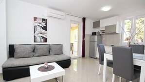 A modern living room with a grey sofa, a small white table, and a kitchen area with a fridge, microwave, and dining table. - Apartment Gorica 1 No.4 - with Pool, 200 m from the Beach (Baska)