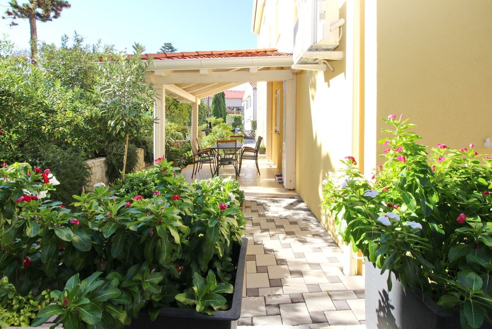 A covered outdoor patio with a tiled floor, surrounded by potted plants and a table with chairs.