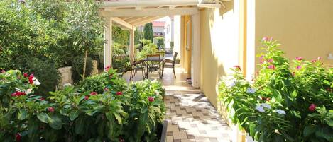 A covered outdoor patio with a tiled floor, surrounded by potted plants and a table with chairs.