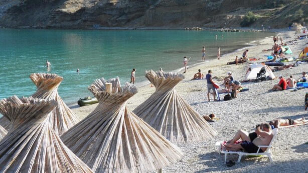 Ein Strand mit Strohdächern, Menschen beim Sonnenbaden und einem klaren türkisfarbenen Meer.