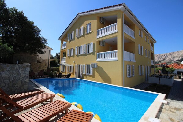 Un hotel de varios pisos con piscina, zona de estar exterior y montañas al fondo.