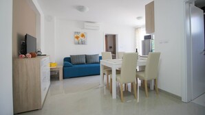 A modern living room with a blue sofa, a dining table with chairs, a kitchen area with a refrigerator, and a TV on a wooden cabinet.
