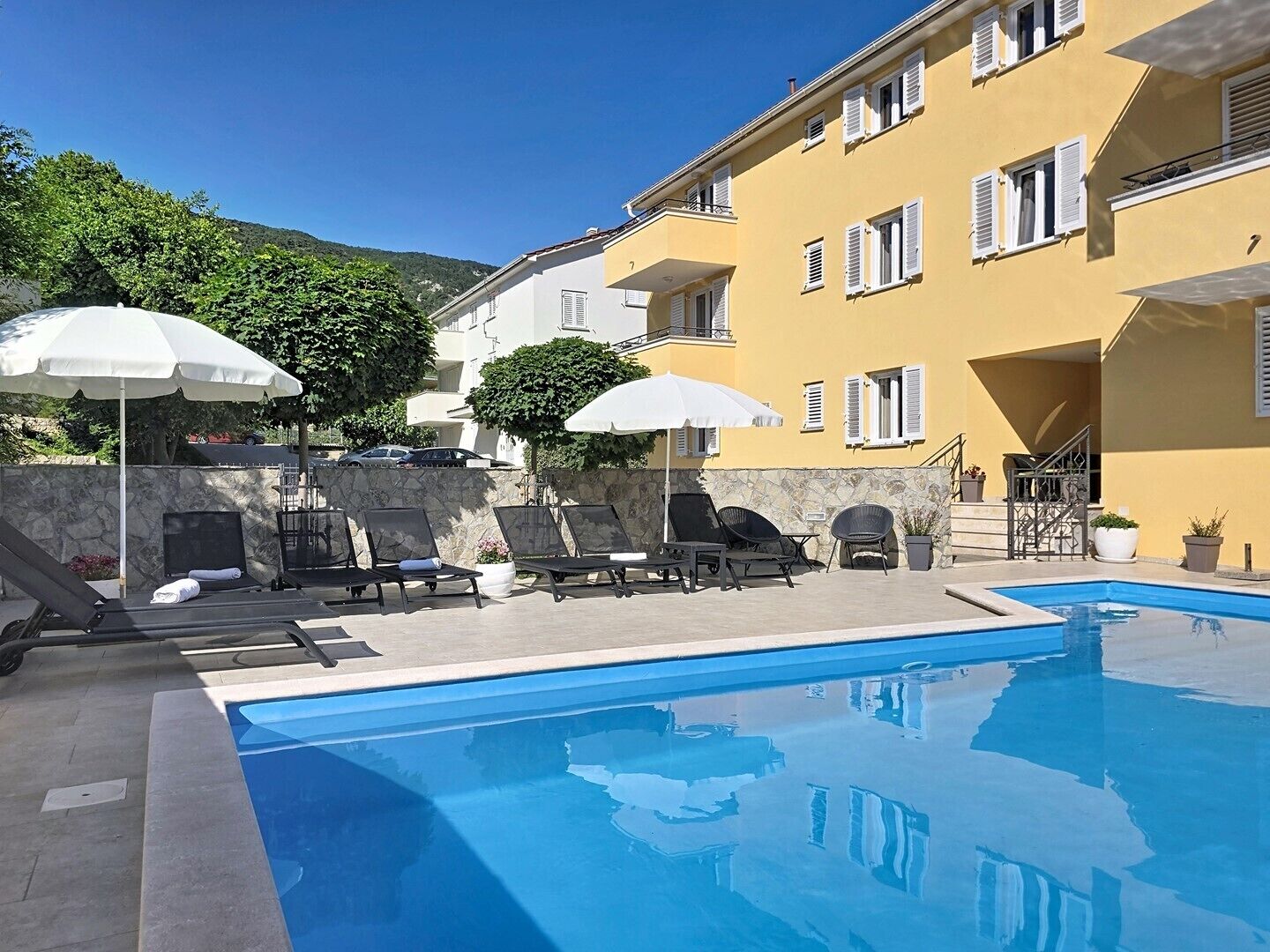 Ein Swimmingpool mit Liegestühlen und Sonnenschirmen, eine Steinmauer und ein gelbes Gebäude mit weißen Fensterläden.
