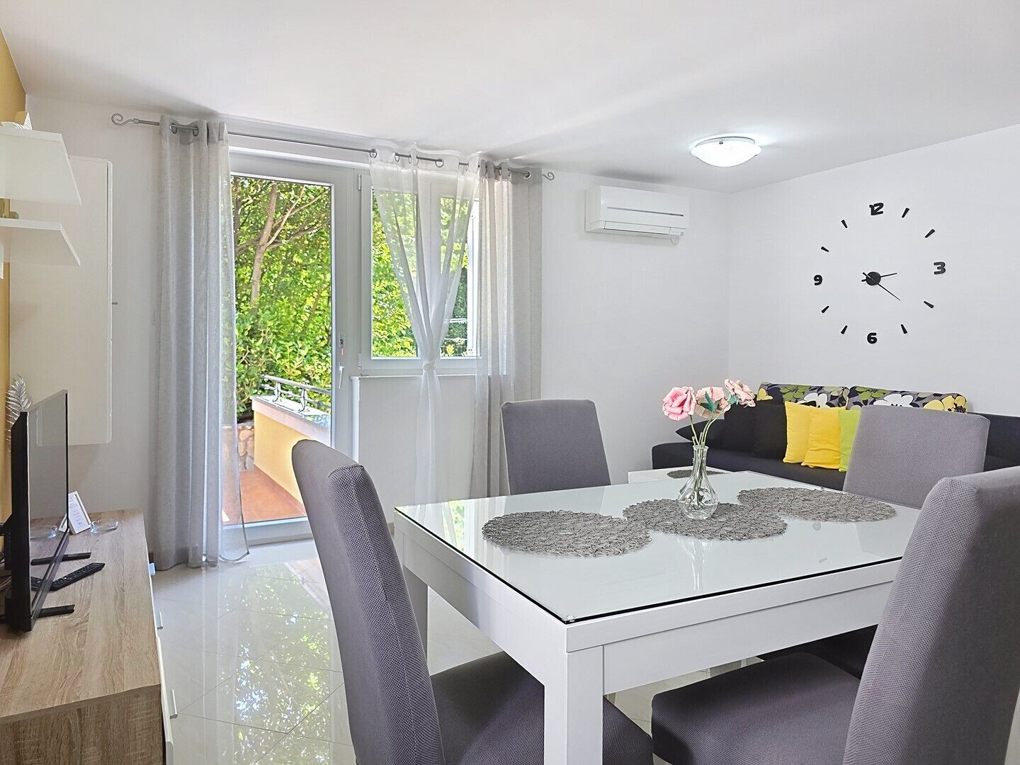 A modern living room with a glass dining table, grey chairs, a sofa with yellow cushions, and a wall clock.