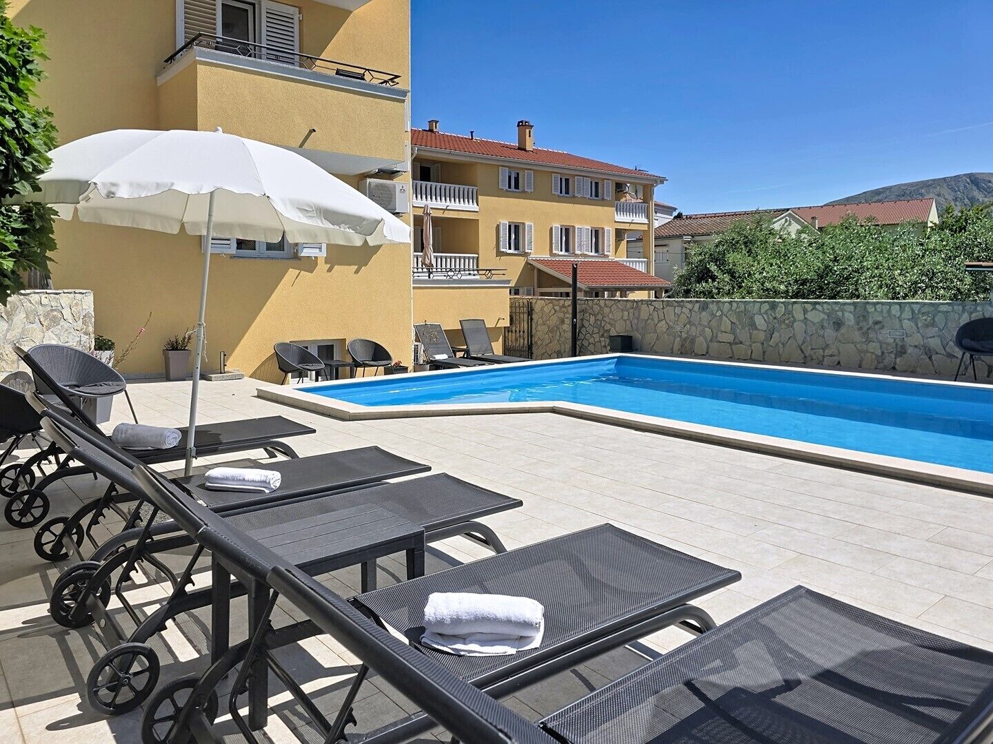 Un área junto a la piscina con sillas de descanso, una sombrilla blanca y un muro de piedra.
