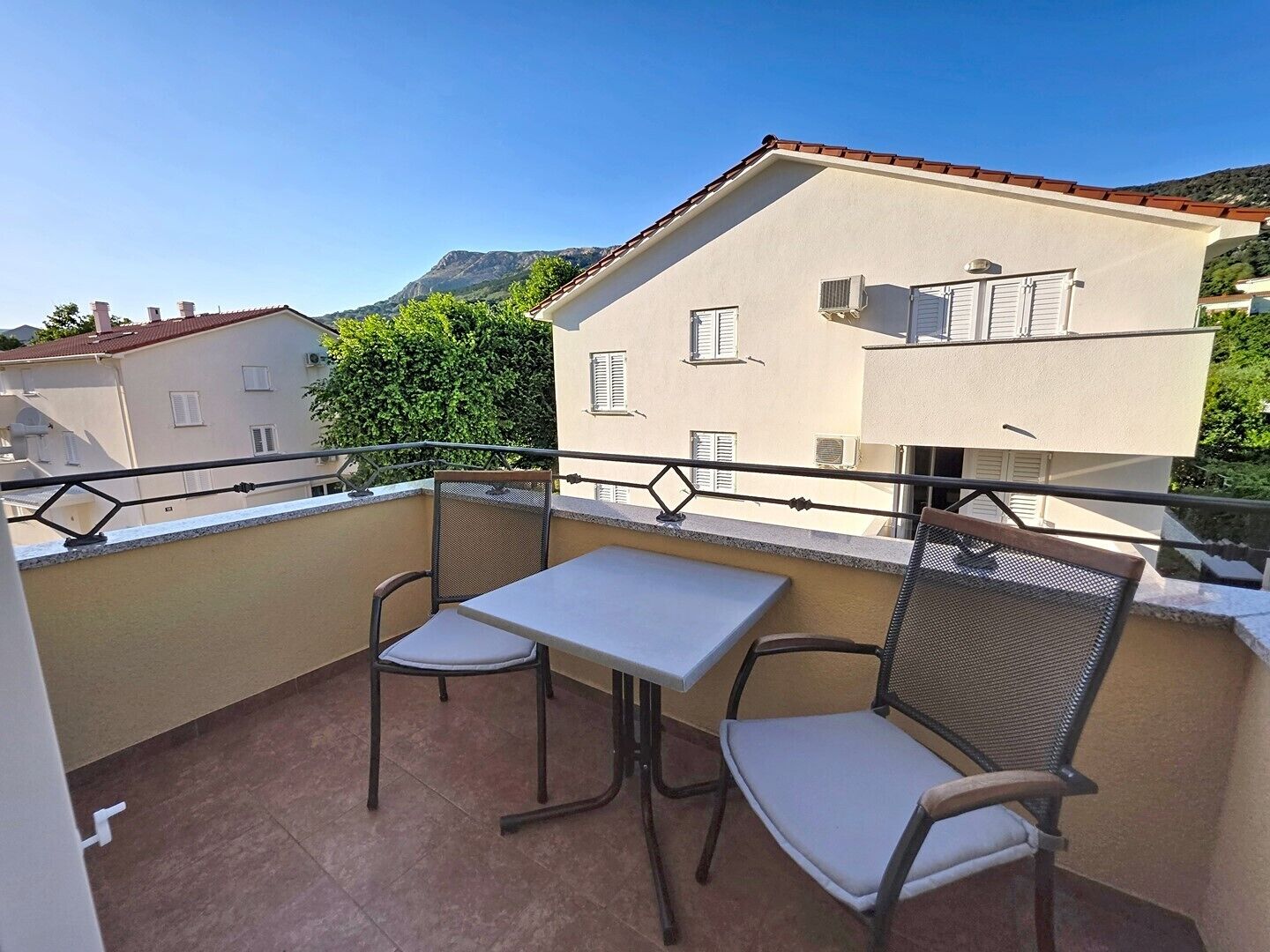 Ein Balkon mit Tisch und zwei Stühlen, Blick auf ein Gebäude mit rotem Dach und ein Berg im Hintergrund.