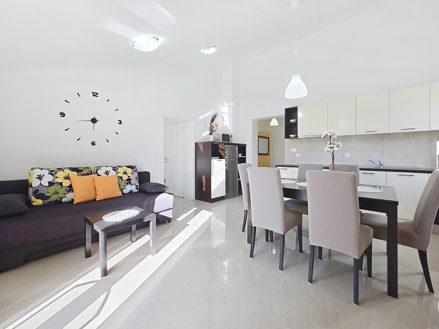 A modern living room with a sofa, a dining table with chairs, a kitchen area, and a wall clock.