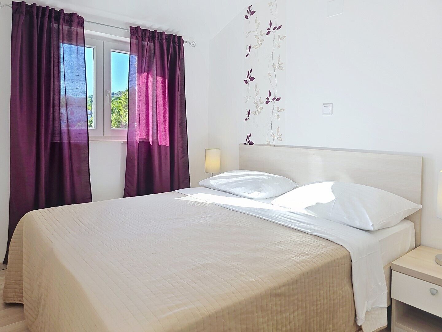 A bed with white linens, a nightstand with a lamp, and a window with purple curtains.